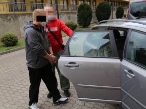 Na zdjęciu widoczny jest zatrzymany mężczyzna z kajdankami na nadgarstkach, którego prowadzi do nieoznakowanego radiowozu nieumundurowany policjant