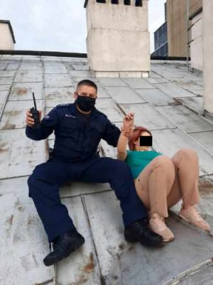Na zdjęciu widoczny jest umundurowany policjant siedzący na dachu budynku i trzymający za rękę poszukiwaną kobietę, która próbowała uciec