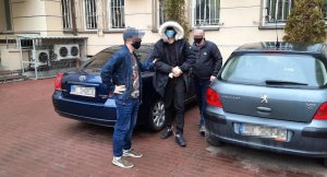 Na zdjęciu widoczny jest zatrzymany mężczyzna prowadzony po parkingu komendy przez nieumundurowanych policjantów